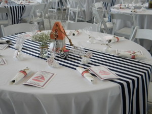 A table set up with a white table cloth and a black and white pin-striped table runner. The table also has programs laid out for the event with goodie bags for the attendees. Additionally there is a flower arrangement, a cup with sugar, and an orange bird feeder sitting on the table.