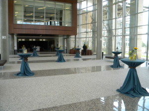 Several bistro tables set up in the AgriLife Center with teal table cloths and flowers on top of the tables. There is also an individual setting up a refreshments table in the back.
