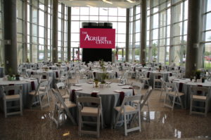 The AgriLife Center set up for an event. Tables are set up with gray table cloths, centerpieces, napkins, and menus. Additionally, each table has a table number. The screen has the AgriLife Center logo displayed.