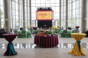 An employee appreciation party held at the AgriLife Center (past event). There are tables set up with green table cloths, bistro tables with a mixture of maroon and yellow table cloths, and food stations set up throughout the room. The screen has the event invitation on display.