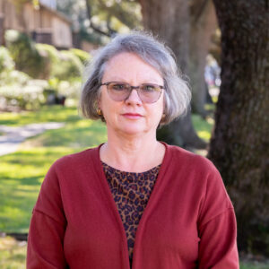 White woman with short grey hair and black glasses. Wearing a red cardigan with a leopard print shirt
